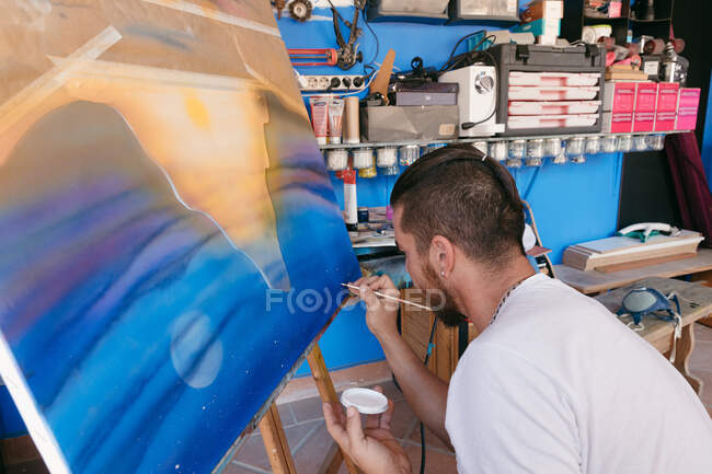 Бородатый человек рисует точки с белым пигментом на холсте с абстрактной картиной во время работы в творческой мастерской — стоковое фото