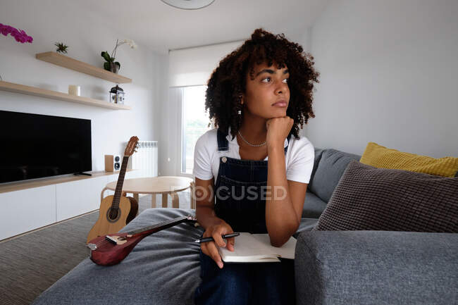 Pensativo músico afroamericano sentado en la cama con mandolina y cuaderno mientras compone la canción y mira hacia otro lado en casa - foto de stock