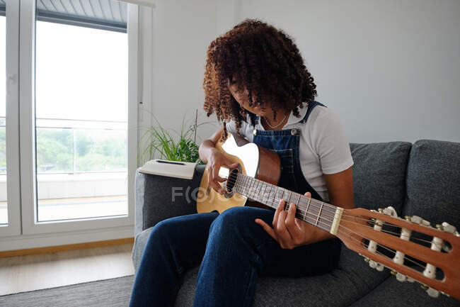 Talented African American female musician playing acoustic guitar while sitting on sofa at home — Stock Photo