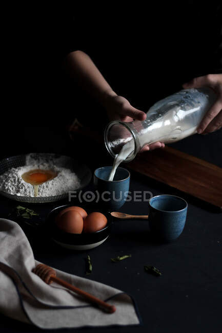 Обрізати руки анонімної жінки під драматичним освітленням готуючи такі інгредієнти, як молоко, борошно та яйця для того, щоб зробити тісто — стокове фото