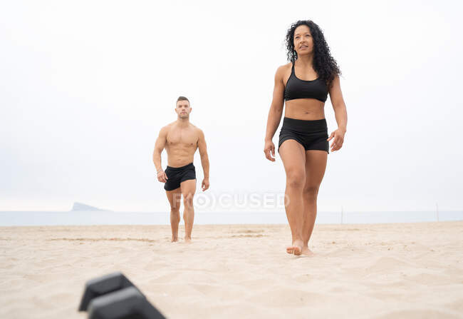 Nivel del suelo de la deportista multirracial y deportista caminando a lo largo de la orilla del mar de arena durante el entrenamiento de fitness en verano - foto de stock