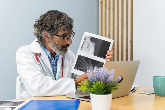 Elderly male medic conducting telemedicine appointment and showing x ray image to patient while using netbook in medical office — Stock Photo