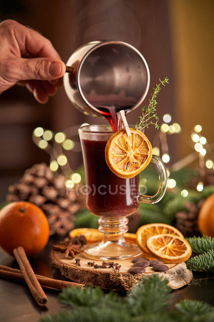 Persona de la cosecha anónima mano que sirve gluhwein o vino caliente de ponche de Navidad en una taza de vidrio con rodajas de naranja secas - foto de stock