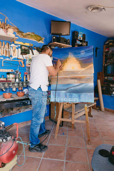 Man in respirator spraying paint on canvas with abstract landscape while working in professional creative studio — Stock Photo