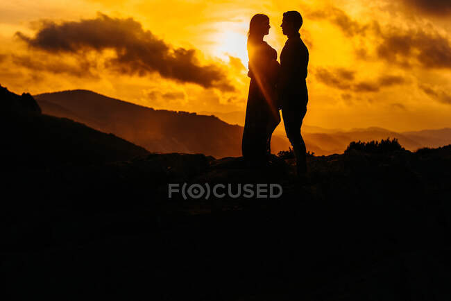 Силуэты с низким углом обзора неузнаваемой пары, ожидающей ребенка, стоящего, глядя друг на друга, держась за руки, любуясь закатом в горах вместе — стоковое фото