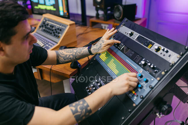 Vista lateral del ingeniero de sonido masculino tatuado de cultivo que trabaja con el panel de control en la placa de audio en el estudio de grabación - foto de stock