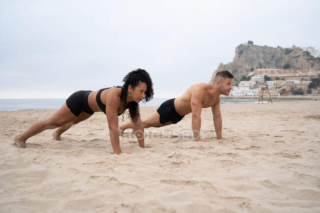 Sportive multiethnic male and female athletes doing push ups during fitness workout on sandy seashore — Stock Photo