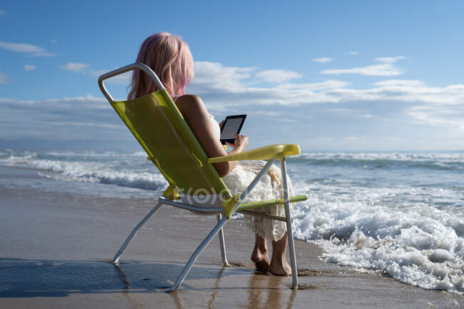 Seitenansicht niedriger Winkel der nicht wiederzuerkennenden Frau mit rosa Haaren, die am Strand chillt, während sie im Sommer auf dem Tablet-PC liest — Stockfoto