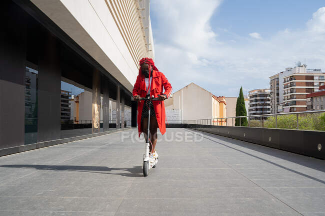 Повний корпус афро-американської самиці в масці з яскравими браїдами їздить електричним скутером вздовж будівлі вежі в вільний час. — стокове фото