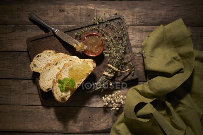 Composición vista superior de delicioso pan casero en rodajas con hierbas aromáticas y mermelada de fruta amarilla dulce en mesa de madera rústica - foto de stock