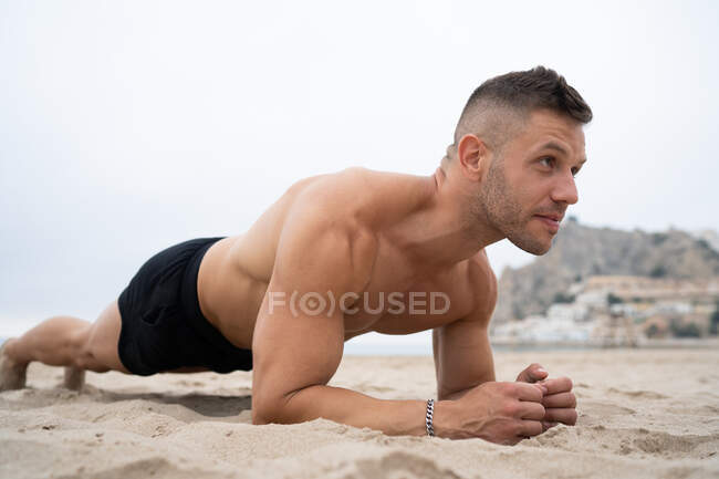 Vista lateral del atleta masculino sin camisa en forma haciendo ejercicio de tablón mientras entrena en la orilla arenosa y mirando hacia otro lado - foto de stock