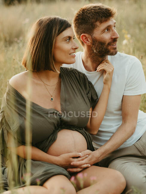 Delighted male hugging of pregnant female with eyes closed while sitting in meadow in countryside looking away — Stock Photo