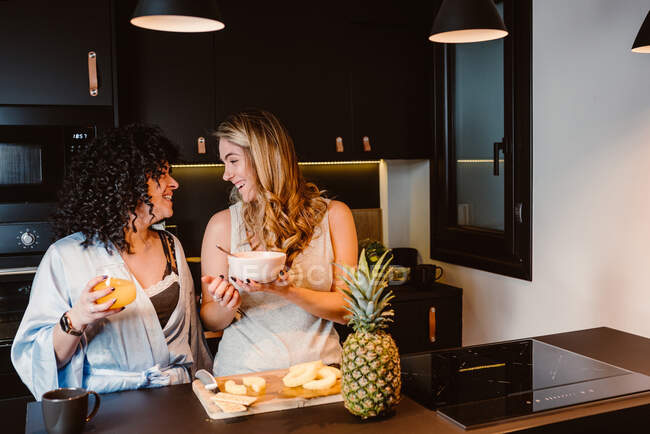 Heiteres lesbisches Paar lacht glücklich, während es Ananas schneidet und Müsli mit Beeren und Joghurt isst — Stockfoto