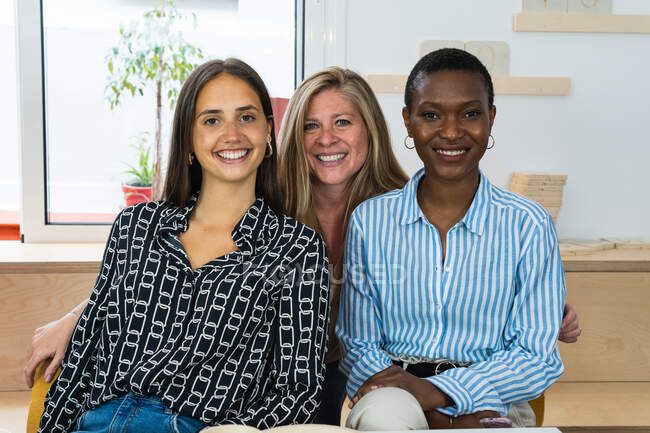 Encantados colegas femeninos multirraciales mirando a la cámara y sonriendo mientras están sentados en el lugar de trabajo juntos - foto de stock