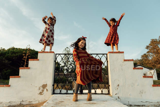 Низький кут повне тіло милих маленьких етнічних дівчат в барвистих сукнях, які розважаються і танцюють на старих кам'яних сходах, насолоджуючись літнім днем разом у сільській місцевості — стокове фото