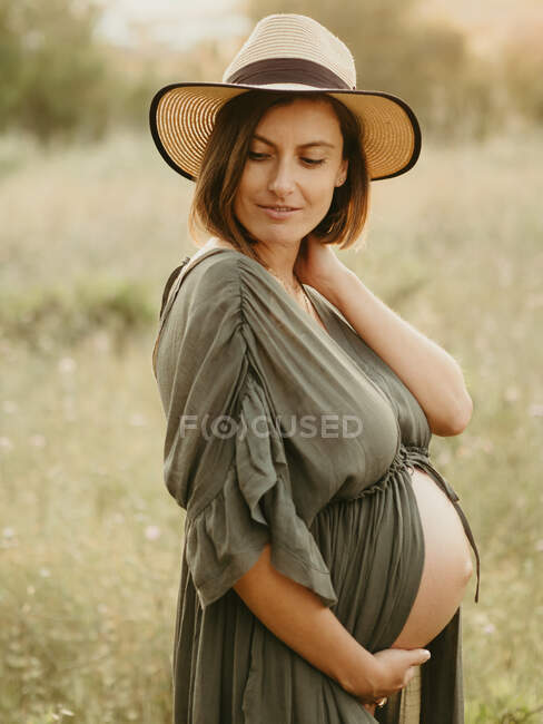 Tranquil pregnant female in dress and straw hat touching tummy while standing in field in countryside at sunset in summer looking away — Stock Photo