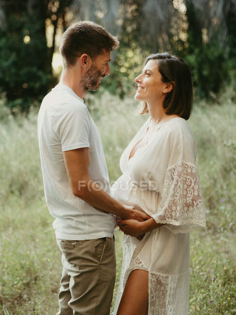 Вид збоку ніжний чоловік тримає руки з захопленою вагітною жінкою, стоячи, дивлячись один на одного в полі в природі — стокове фото