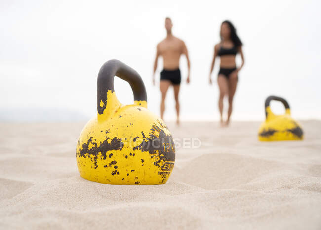 Ground level of yellow shabby metal kettlebell placed on sandy beach on background of blurred sportsman and sportswoman — Stock Photo