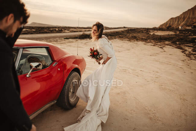 Unrecognizable man in suit standing against red sports car and cheerful bride at sunset in Bardenas Reales Natural Park in Navarra, Spain — Stock Photo