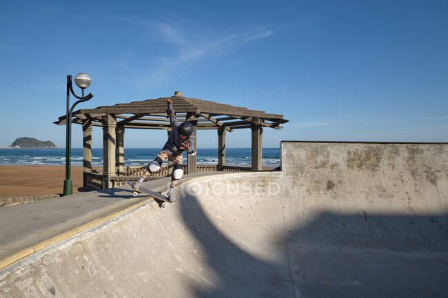 Unrecognizable teen boy in protective helmet riding skateboard in skate park on sunny day on seashore — Stock Photo