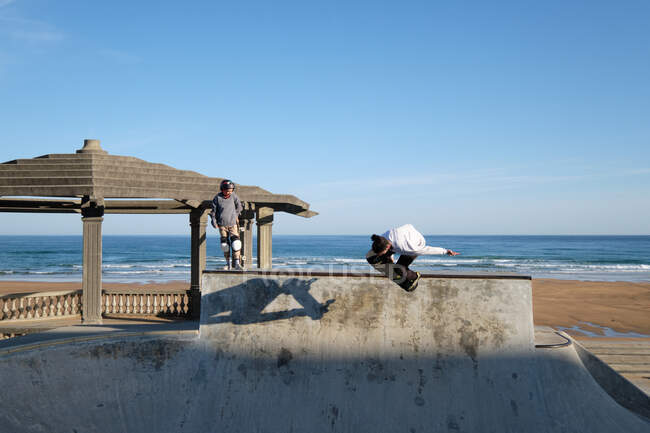 Pattinatori attivi cavalcando skateboard e mostrando trucchi in skate park al mare in estate — Foto stock
