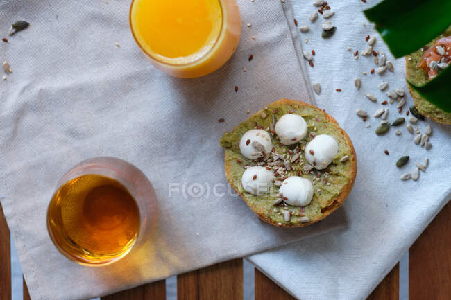From above of yummy toast with avocado and mozzarella balls decorated with flax and sunflower seeds and served on napkin near glasses of fresh orange juice and herbal tea — Stock Photo