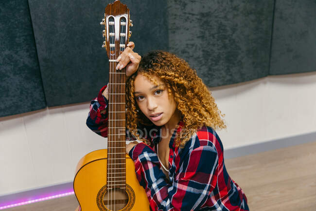 Joven guitarrista étnica femenina con el pelo rizado apoyado en la guitarra acústica mientras mira a la cámara - foto de stock