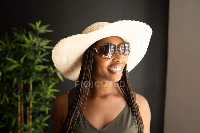 Heureuse femme afro-américaine branchée portant un chapeau de paille et des lunettes de soleil regardant loin dans la chambre à la maison — Photo de stock