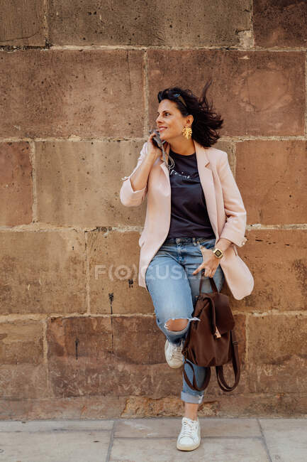 Mujer hispana adulta en pantalones vaqueros rasgados casuales de moda y blazer con mochila sobre el hombro riendo felizmente durante la conversación telefónica mientras está de pie en la pared urbana - foto de stock