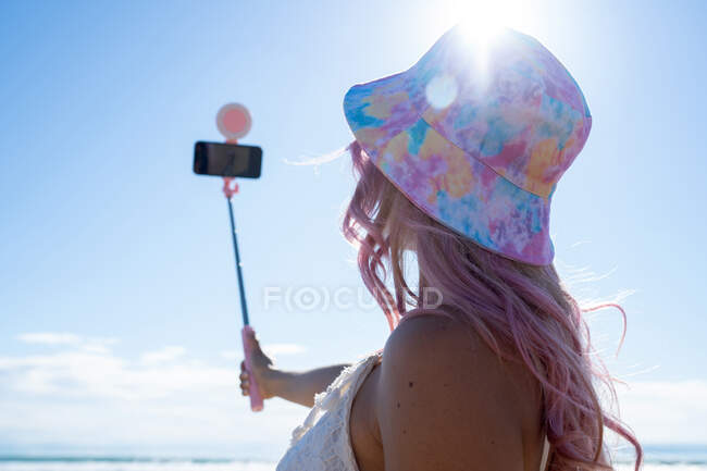 Неузнаваемая женщина с розовыми волосами и в летнем наряде делает самоснимок на смартфоне на селфи-палочке, стоя в солнечный день на берегу моря — стоковое фото