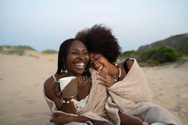 Happy young black female best friends with mugs of hot drinks sitting close wrapped in warm blanket and having fun during summer evening on sandy beach — Stock Photo