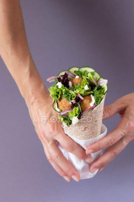 Cultivado irreconocible persona manos sosteniendo envoltura de falafel vegano sobre fondo púrpura colorido - foto de stock