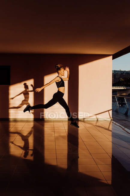 Joven atlética caucásica haciendo ejercicio al atardecer practicando saltos, sombras y luz sobre fondo - foto de stock