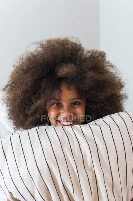 Fröhliches afroamerikanisches Teenie-Mädchen mit Afro-Frisur versteckt sich zu Hause hinter einem Kissen und blickt in die Kamera — Stockfoto