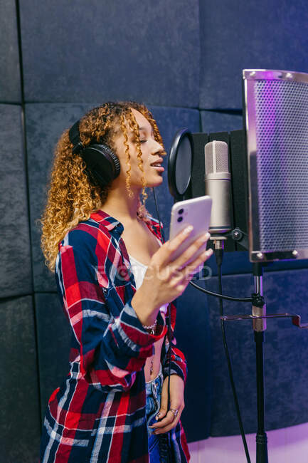 Cantora afro-americana alegre em fones de ouvido com smartphone tocando música contra escudo sonoro em estúdio de gravação — Fotografia de Stock
