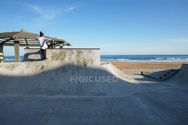 Irreconocible adolescente en protector casco de montar monopatín en skate park en día soleado en la orilla del mar - foto de stock