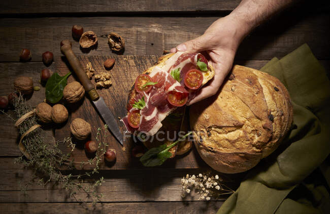 Vista superior da pessoa sem rosto cultura segurando delicioso sanduíche caseiro feito com pão artesanal com tomate cereja e presunto com ervas e nozes — Fotografia de Stock
