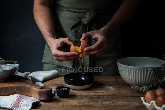 Homme méconnaissable dans tablier casser oeuf cru sur bol tout en préparant la pâtisserie sur la table de bois près des ustensiles de cuisine — Photo de stock