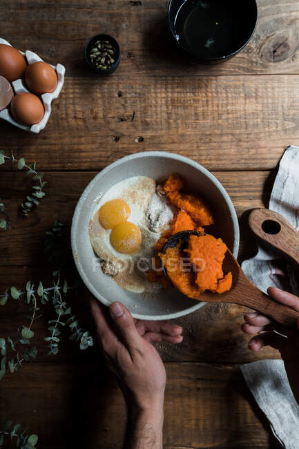 Зверху невизначений чоловічий кухар використовує дерев'яну ложку, щоб змішати гарбузове пюре з яйцями та борошном в мисці, готуючи пиріг на дерев'яному столі біля обробної дошки та рушника — стокове фото
