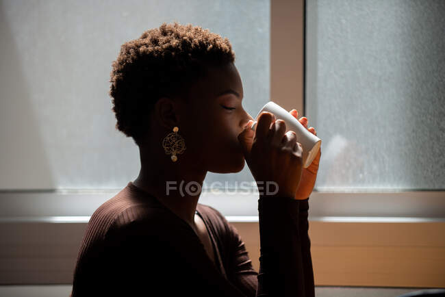 Vista lateral de la hembra afroamericana con cabello corto bebiendo bebida refrescante de la taza en casa por la mañana - foto de stock
