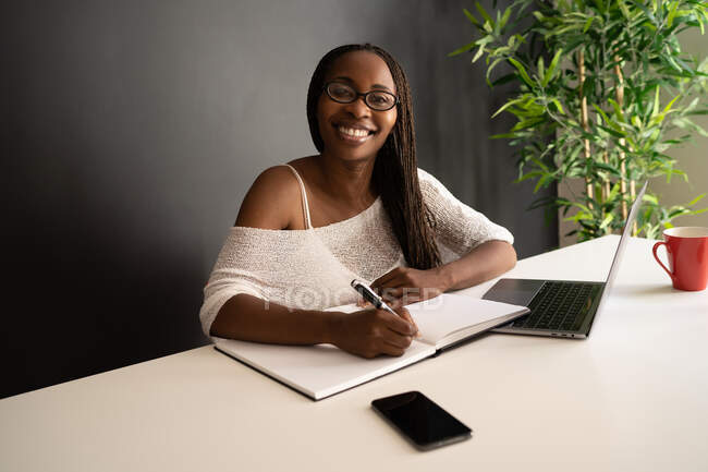 Content black female freelancer writing notes in notebook while sitting at table with laptop and working on business project in modern home office — Stock Photo