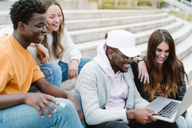 Разнообразные молодые друзья широко улыбаются во время просмотра видео на нетбуке вместе, сидя на бетонной лестнице — стоковое фото