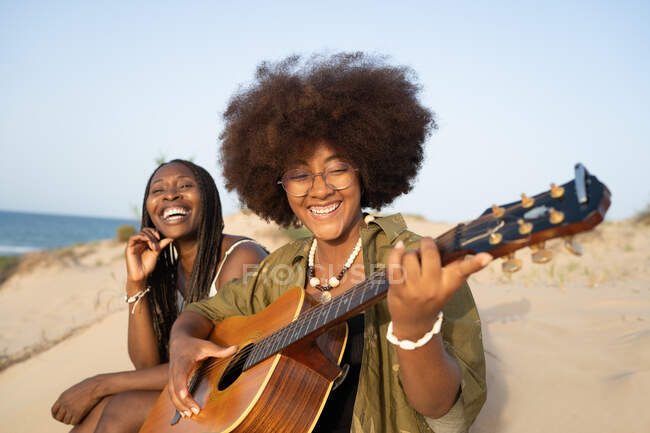 Felices amigas afroamericanas jóvenes tocando la guitarra mientras están sentadas juntas en la orilla del mar y disfrutando de las vacaciones de verano. - foto de stock