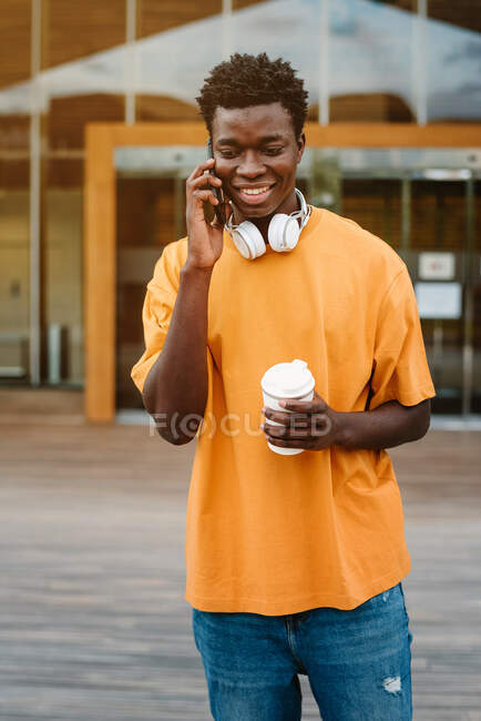 Sonriente hombre afroamericano con taza de café riendo felizmente mientras habla por teléfono móvil y mirando hacia otro lado - foto de stock