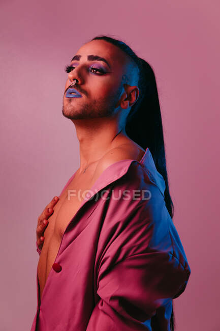 Portrait de femme barbu transgenre glamour dans un maquillage sophistiqué posant sur fond rose au studio détournant les yeux — Photo de stock
