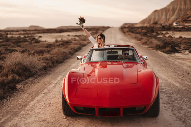 Bride raising arm with bouquet while riding red sports car with groom during trip through Bardenas Reales Natural Park in Navarra, Spain — Stock Photo