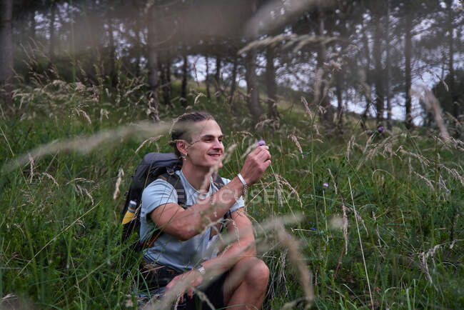 Caminhante masculino alegre sentado no campo com flor e curtindo trekking na floresta no verão — Fotografia de Stock