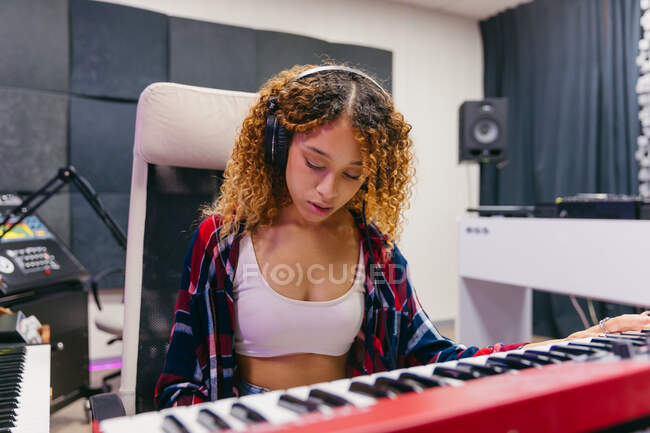 Focus African American female singer in headset playing synthesizer while performing song into mic in recording studio — Stock Photo