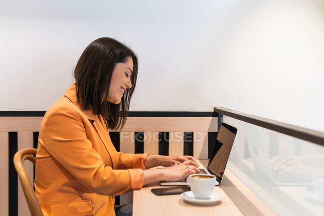 Side view of happy female freelancer browsing netbook while sitting at table with cup of coffee and working remotely in cafe — Stock Photo