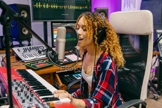 Vista lateral da cantora afro-americana sorridente no fone de ouvido tocando sintetizador enquanto executa música no microfone no estúdio de gravação — Fotografia de Stock